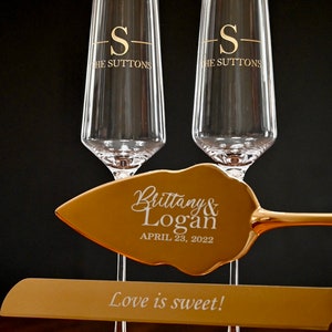 May include: Two gold champagne flutes with the inscription "S- THE SUTTONS" on each. A gold cake server with the inscription "Brittany & Logan APRIL 23, 2022" and a gold base with the inscription "Love is sweet!"