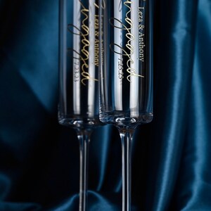 May include: Two clear glass champagne flutes with gold lettering that reads "Engaged" and "Leza & Anthony 7.23.23". The flutes are on a dark blue satin background.