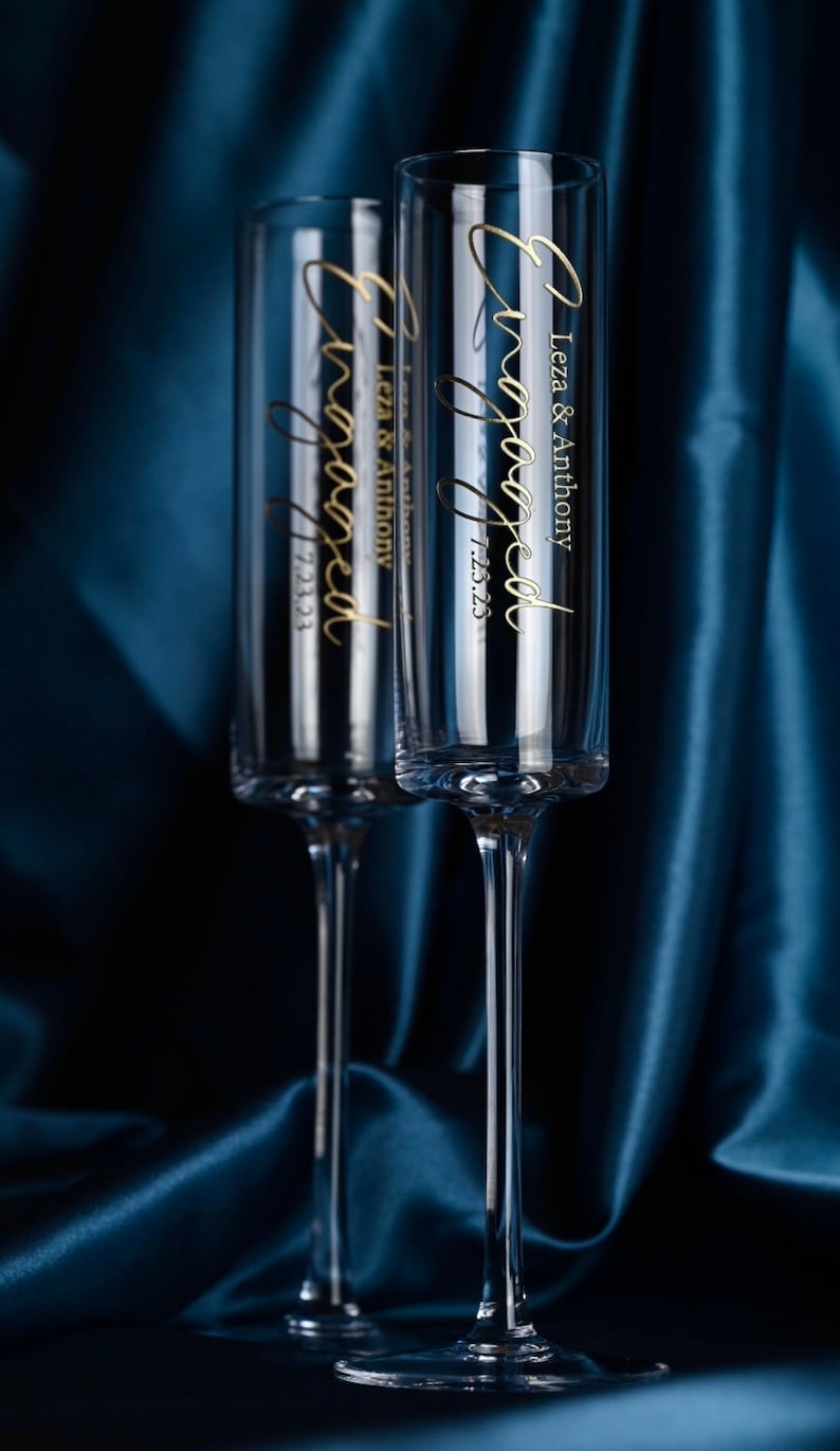 May include: Two clear glass champagne flutes with gold lettering that reads "Engaged" and "Leza & Anthony 7.23.23". The flutes are on a dark blue satin background.