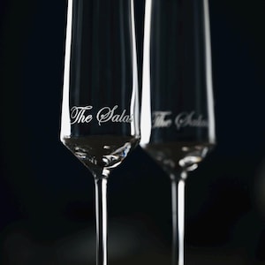Puede incluir: Dos copas de champ&aacute;n transparentes con el texto "The Salas" grabado en el cristal. Las copas tienen un dise&ntilde;o alto y delgado con un tallo largo y una base ancha. El fondo es oscuro.