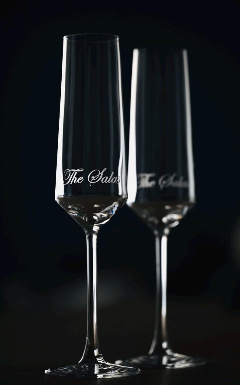 Puede incluir: Dos copas de champ&aacute;n transparentes con el texto "The Salas" grabado en el cristal. Las copas tienen un dise&ntilde;o alto y delgado con un tallo largo y una base ancha. El fondo es oscuro.