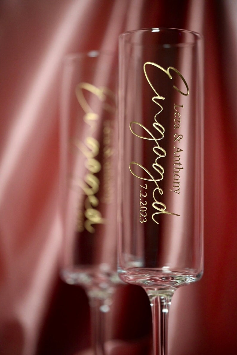 May include: Two clear champagne flutes with gold lettering that reads "Engaged" and "Leza & Anthony 7.2.2023". The flutes are in front of a red and pink satin background.