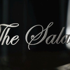 Puede incluir: Primer plano de un vaso con las palabras "The Salas" grabadas en una elegante escritura plateada. El texto est&aacute; centrado sobre un fondo oscuro, con los bordes del vaso visibles.