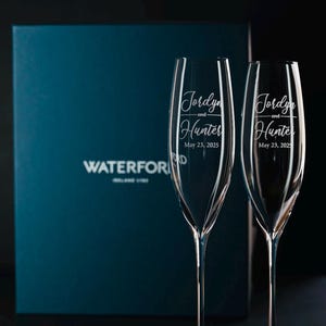 May include: Two clear glass champagne flutes with slender stems and bowls. Each glass is etched with the names Jordyn and Hunter, the word and, and the date May 23, 2025. A dark blue box with the white text WATERFORD IRELAND 1783 is visible behind.