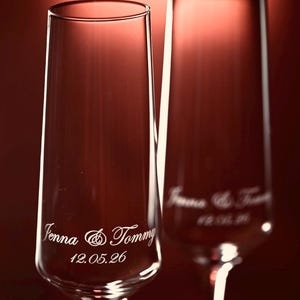 Puede incluir: Dos copas de champ&aacute;n transparentes con grabado personalizado. El texto en la primera copa dice "Jenna & Tommy" y "12.05.26". La segunda copa tiene un texto similar. Las copas est&aacute;n sobre un fondo rojo.