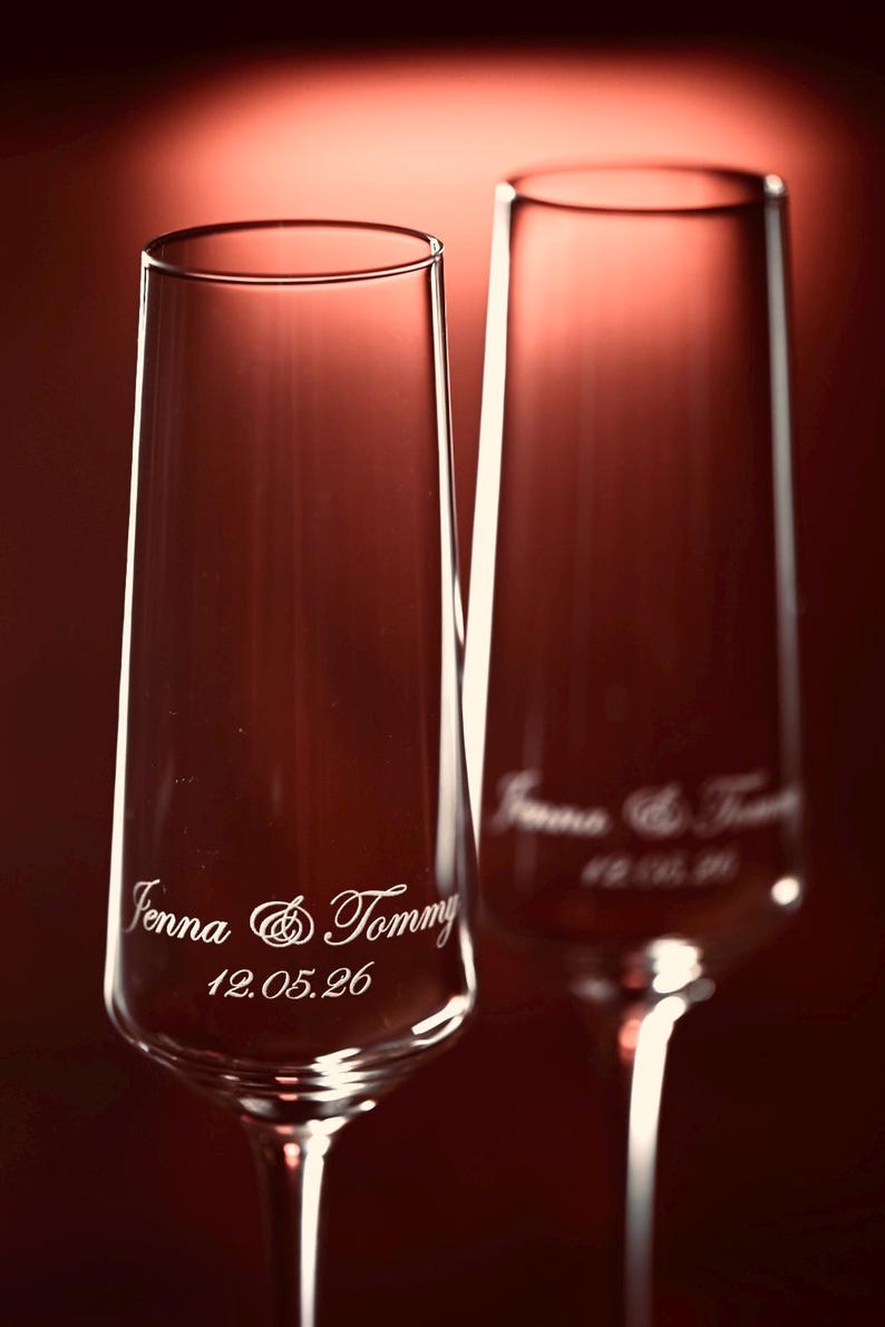 Puede incluir: Dos copas de champ&aacute;n transparentes con grabado personalizado. El texto en la primera copa dice "Jenna & Tommy" y "12.05.26". La segunda copa tiene un texto similar. Las copas est&aacute;n sobre un fondo rojo.