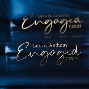 May include: Two clear glass champagne flutes with gold lettering that reads "Leza & Anthony Engaged 7.23.23". The flutes are on a dark blue satin fabric.