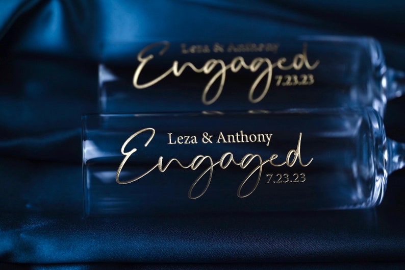 May include: Two clear glass champagne flutes with gold lettering that reads "Leza & Anthony Engaged 7.23.23". The flutes are on a dark blue satin fabric.