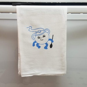 Może przedstawiać: Biały ręcznik kuchenny z niebieską kreskówkową ilustracją czajnika z uśmiechającą się twarzą i ramionami trzymającymi łyżkę.