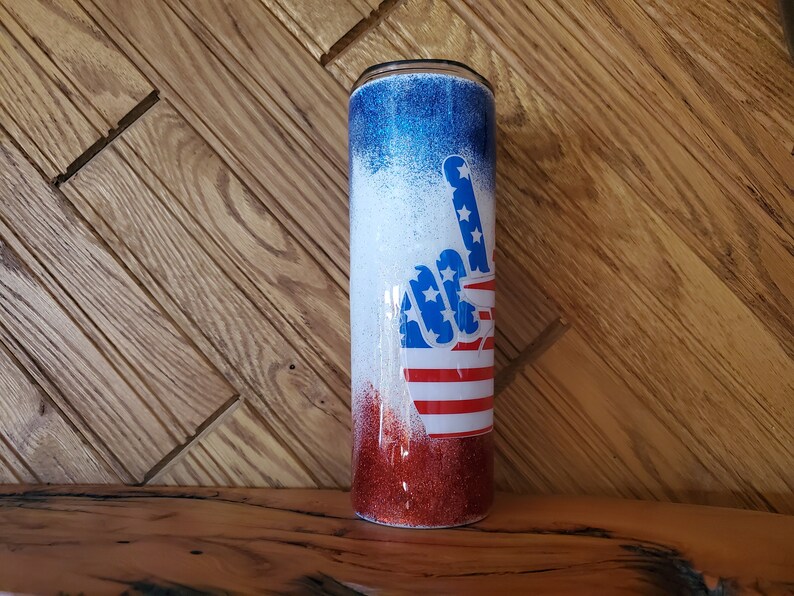 May include: A red, white, and blue glitter tumbler with a peace sign design. The peace sign is made up of the American flag.