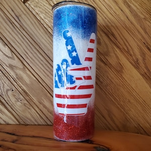 May include: A red, white, and blue glitter tumbler with a peace sign design. The peace sign is made up of the American flag.