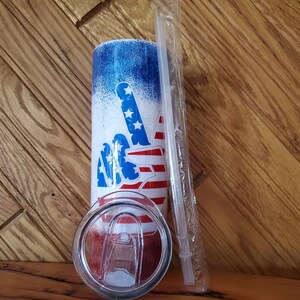 May include: A white tumbler with a blue and red peace sign design. The tumbler has a clear lid and a clear straw. The design features a peace sign with the American flag colors.