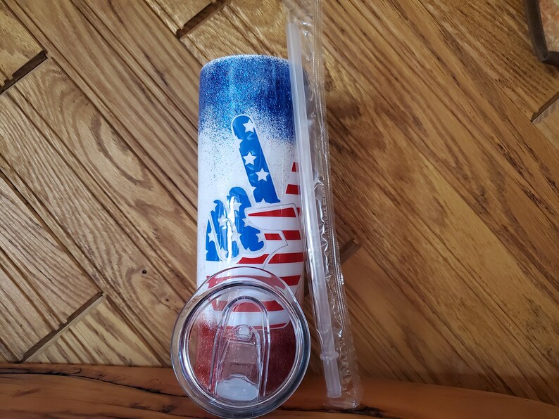 May include: A white tumbler with a blue and red peace sign design. The tumbler has a clear lid and a clear straw. The design features a peace sign with the American flag colors.