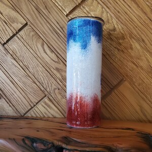 May include: A red, white, and blue ombre glitter tumbler with a silver lid. The tumbler is on a wooden surface.