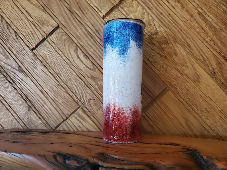 May include: A red, white, and blue ombre glitter tumbler with a silver lid. The tumbler is on a wooden surface.