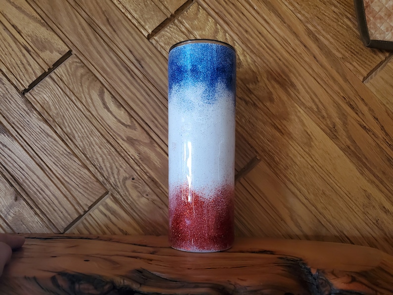 May include: A red, white, and blue ombre glitter tumbler. The tumbler has a stainless steel exterior and a clear lid.