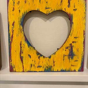 May include: A square wooden picture frame with a heart-shaped cutout. The frame is painted with a textured yellow paint, with hints of blue and purple peeking through. The frame is sitting on a white shelf.
