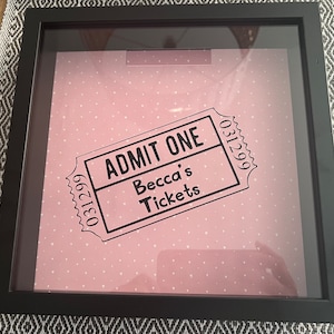 May include: A black shadow box frame with a pink background with white hearts. A black and white ticket with the text "ADMIT ONE" and "Becca's Tickets" is displayed in the frame.