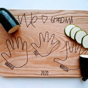 May include: A wooden cutting board with three handprints and the names Frankie, Paul, and Waylon. The board also has the text "We ♥ Grandma" and "2020". There are two zucchini halves and several zucchini slices on the board.