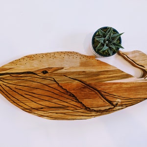 May include: A wooden cutting board shaped like a whale with a burnt design. The whale has a detailed eye and a small succulent plant in a pot is sitting on top of the whale's tail.
