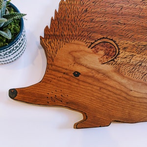 May include: A wooden cutting board with a detailed woodburned design of a hedgehog. The hedgehog is facing left and has a brown body with black eyes and a black nose.