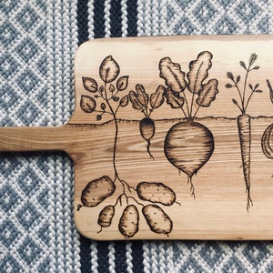 May include: A wooden cutting board with a hand-painted design of various vegetables, including potatoes, carrots, onions, and beets. The board has a handle on the left side.