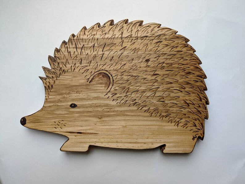 May include: A wooden cutting board shaped like a hedgehog with a detailed burned design. The board is light brown with a natural wood grain.
