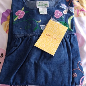 May include: A blue denim dress with embroidered flowers and a dragonfly. The dress has a round neckline and a gathered skirt. The dress has a tag that says "QVC style".