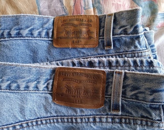 May include: Close-up of two pairs of light blue denim jeans. Each pair features a brown leather patch with the Levi Strauss & Co. logo and "545" printed on it. The jeans are folded, showing the waistband and belt loops.