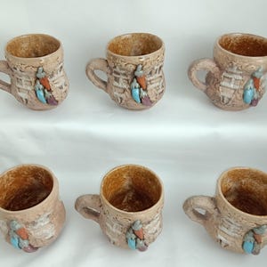 May include: Six ceramic mugs with handles, featuring a textured, beige exterior and a brown interior. Each mug is adorned with a colorful feather design in shades of red, blue, and purple. The mugs are arranged on a white surface.