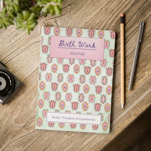 May include: A journal with a light green cover featuring a repeating pattern of pink and orange floral designs. The journal has a pink rectangular label with the words "Birth Work Journal" and "Notes, Thoughts, & Experiences". A pencil and pen are next to the journal.