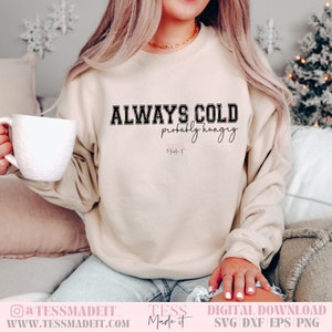 以下が含まれることがあります： ベージュのトレーナーに、「ALWAYS COLD probably hungry」という黒い文字が書かれています。文字は大学風のフォントです。トレーナーは、ソファに座っている人が着用しています。トレーナーには、「TESS Made it」という文字も書かれています。