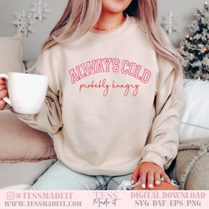 May include: A beige sweatshirt with the text "ALWAYS COLD probably hungry" in pink lettering. The sweatshirt is being worn by a person holding a white mug.