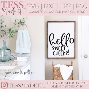 Puede incluir: Un cartel blanco con texto negro que dice "hello sweet cheeks!" colgado en una pared blanca en un ba&ntilde;o. El cartel est&aacute; enmarcado en madera. Hay una toalla blanca colgada en un toallero y una cesta de mimbre con una toalla blanca dentro.