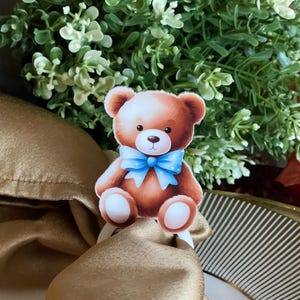 May include: A brown teddy bear with a blue bow tie is attached to a white napkin ring. The ring is on a gold napkin.