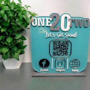 May include: A turquoise blue acrylic sign with the words "ONE20TWO Let's Get Social" in white and gray lettering. The sign features a QR code, Facebook, Instagram, and Website icons.