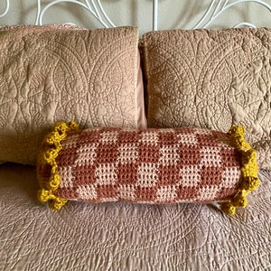 May include: A brown and white checkered crochet bolster pillow with yellow crocheted trim. The pillow is on a bed with a light brown quilted bedspread.