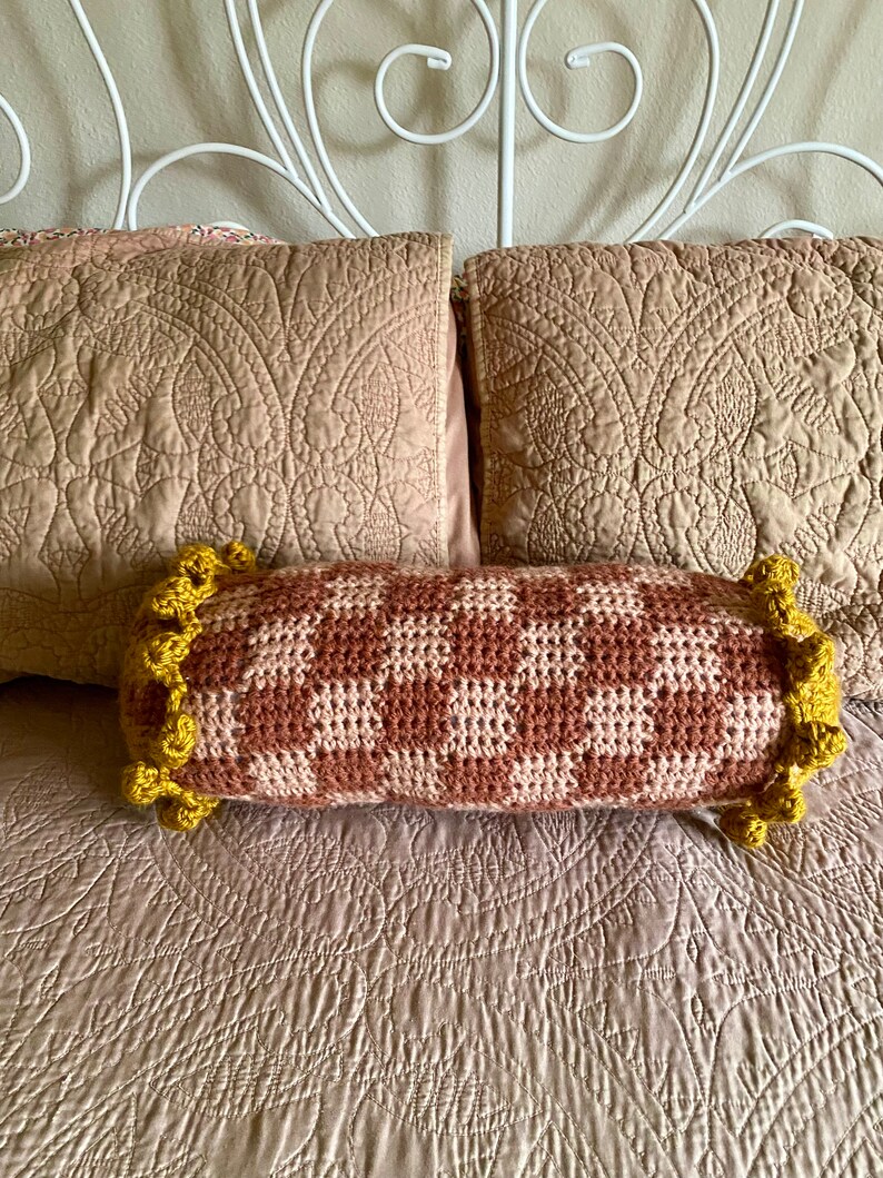 May include: A brown and white checkered crochet bolster pillow with yellow crocheted trim. The pillow is on a bed with a light brown quilted bedspread.