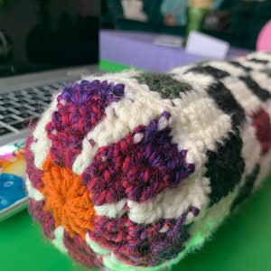 May include: Close-up of a crocheted pillow with a colorful geometric pattern. The pillow is made with white, orange, purple, red, green, and black yarn.
