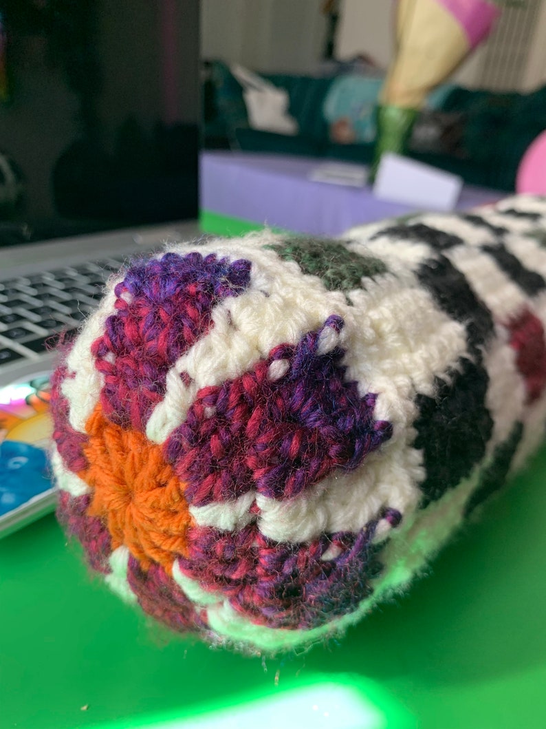 May include: Close-up of a crocheted pillow with a colorful geometric pattern. The pillow is made with white, orange, purple, red, green, and black yarn.