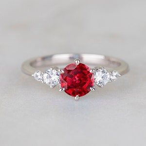 May include: A silver ring with a red gemstone surrounded by smaller white gemstones.