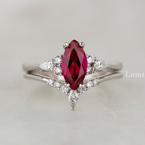 Könnte beinhalten: Ein silberner Ring mit einem rubinroten Marquise-Schliff-Stein, der von kleineren weißen Steinen umgeben ist. Der Ring hat ein filigranes, kunstvolles Design und ist auf einem weißen Hintergrund platziert. 'Lumiel Jewelry' ist in der rechten unteren Ecke des Bildes gedruckt.