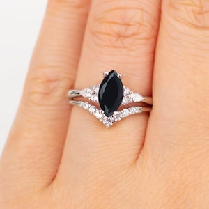 Może przedstawiać: Srebrny pierścionek z czarnym, markizowym kamieniem szlachetnym otoczonym mniejszymi, przezroczystymi kamieniami szlachetnymi. Pierścionek jest noszony na palcu.