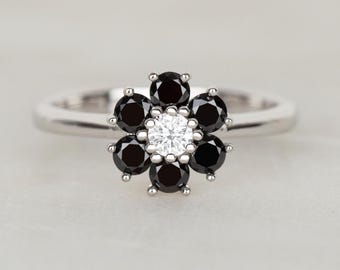 May include: A silver ring featuring a flower design. The flower's petals are formed by black gemstones surrounding a central white gemstone. The ring is set against a soft, neutral background, highlighting the jewelry's elegant design.