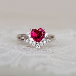K&ouml;nnte beinhalten: Ein Herz-f&ouml;rmiger Rubinring mit einem silbernen Band und kleineren Diamanten, die in einem V-f&ouml;rmigen Band gefasst sind.