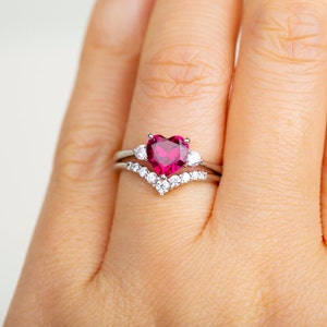 K&ouml;nnte beinhalten: Ein silberner Ring mit einem gro&szlig;en, herzf&ouml;rmigen Rubin, der von kleineren, klaren Edelsteinen umgeben ist.