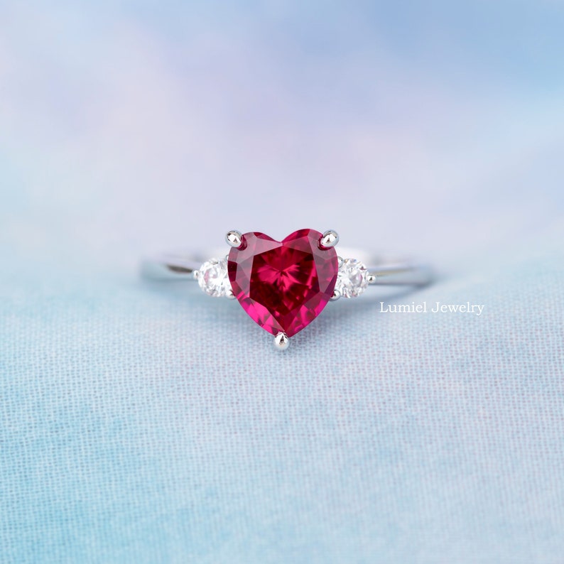 K&ouml;nnte beinhalten: Ein silberner Ring mit einem gro&szlig;en herzf&ouml;rmigen Rubin-Edelstein und zwei kleineren Diamant-Akzenten. Der Ring liegt auf einem hellblauen Stoffhintergrund.