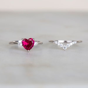 K&ouml;nnte beinhalten: Ein Herz-f&ouml;rmiger Rubin-Verlobungsring mit zwei kleineren Diamanten auf jeder Seite des Mittelst&uuml;cks. Der Ring ist in Silber gefasst und wird mit einem passenden Silberband mit einer Reihe von Diamanten in V-Form kombiniert.