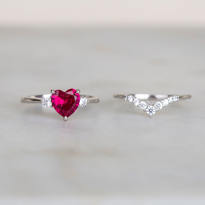 K&ouml;nnte beinhalten: Ein Herz-f&ouml;rmiger Rubin-Verlobungsring mit zwei kleineren Diamanten auf jeder Seite des Mittelst&uuml;cks. Der Ring ist in Silber gefasst und wird mit einem passenden Silberband mit einer Reihe von Diamanten in V-Form kombiniert.