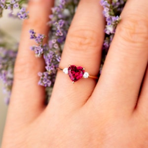 K&ouml;nnte beinhalten: Ein Ring mit einem herzf&ouml;rmigen Rubin und zwei kleineren Diamanten auf einem goldenen Band. Der Ring befindet sich an einem Finger mit neutralem Nagellack.